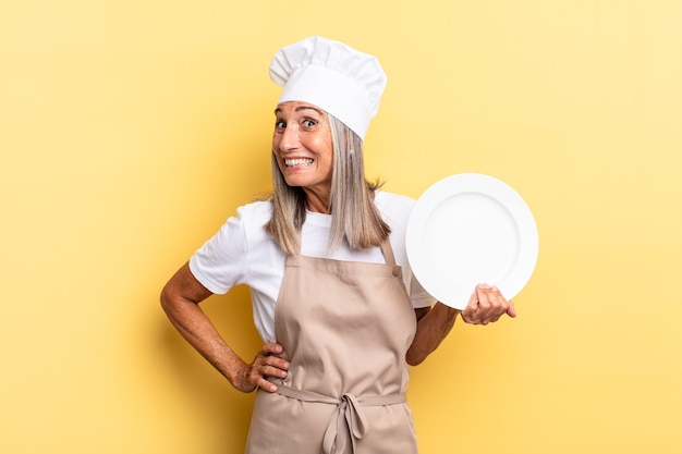 Middle age chef woman shrugging, feeling confused and uncertain, doubting with arms crossed and puzzled look and holding a dish