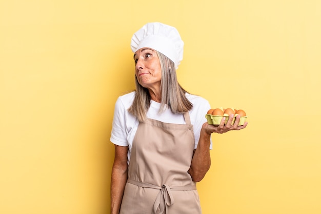 Middle age chef woman shrugging, feeling confused and uncertain, doubting with arms crossed and puzzled look holding an eggs box