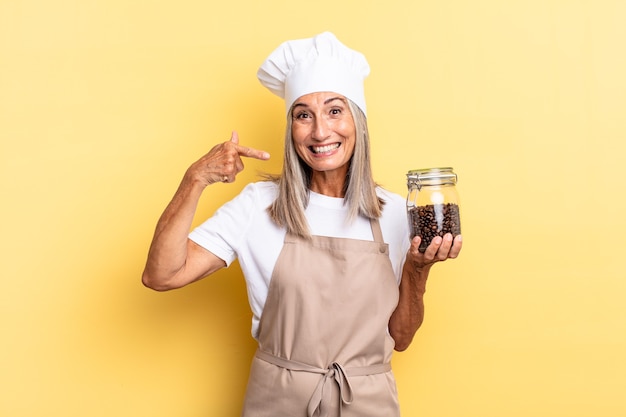 Middle age chef woman smiling confidently pointing to own broad smile, positive, relaxed, satisfied attitude holding coffee beans