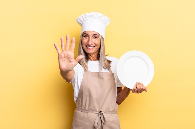 Middle age chef woman smiling and looking friendly, showing number five or fifth with hand forward, counting down and holding a dish