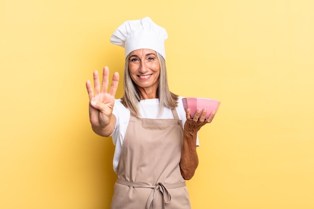 Женщина-шеф-повар средних лет улыбается и выглядит дружелюбно, показывая номер четыре или четвертый рукой вперед, считая в обратном порядке и держа пустую кастрюлю