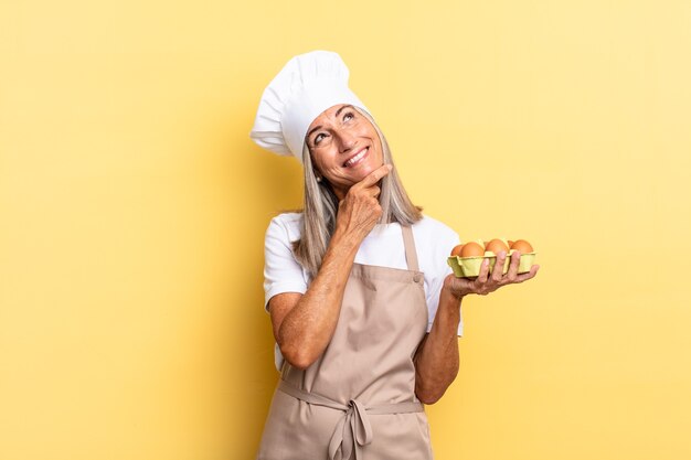 Middle age chef woman smiling with a happy, confident\
expression with hand on chin, wondering and looking to the side\
holding an eggs box