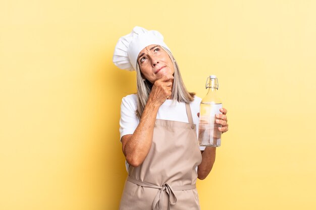 Middle age chef woman thinking, feeling doubtful and confused, with different options, wondering which decision to make with a water bottle