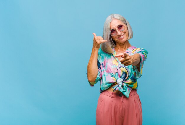 Donna fredda di mezza età che sorride allegramente e punta alla telecamera mentre si effettua una chiamata in seguito gesto, parlando al telefono