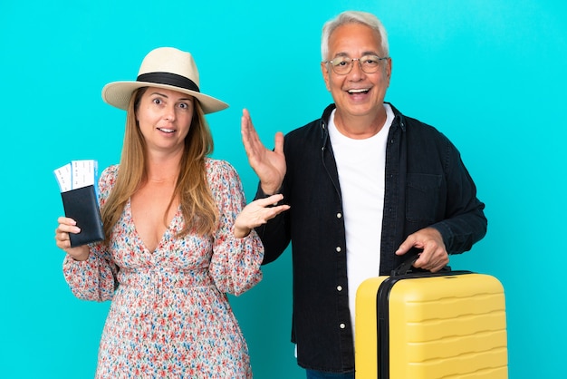 Middle age couple going to travel and holding a suitcase isolated on blue background with surprise and shocked facial expression