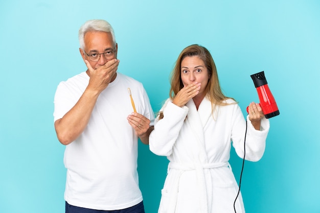 Middle age couple holding dryer and toothbrush isolated on blue background covering mouth with hands for saying something inappropriate