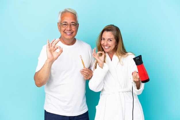 Middle age couple holding dryer and toothbrush isolated on blue background showing an ok sign with fingers