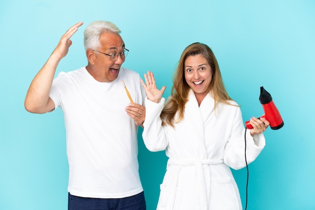 Middle age couple holding dryer and toothbrush isolated on blue background with surprise and shocked facial expression