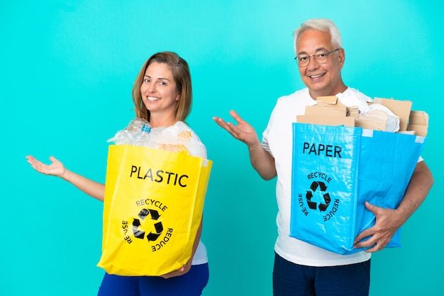 Middle age couple holding a recycling bags full of paper and plastic isolated on white background extending hands to the side for inviting to come