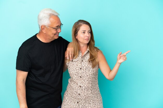 Пара среднего возраста, изолированные на синем фоне, указывая в сторону, чтобы представить продукт