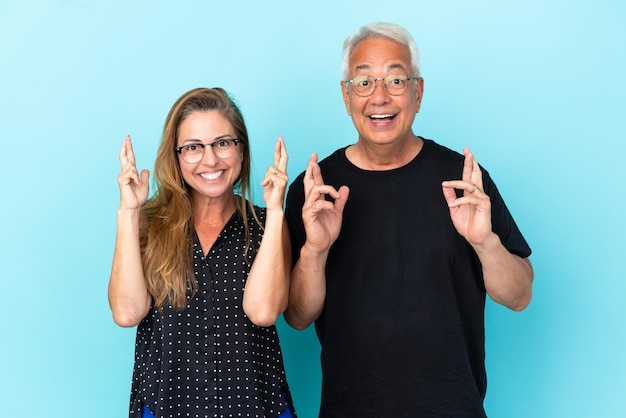 Middle age couple isolated on blue background with fingers crossing