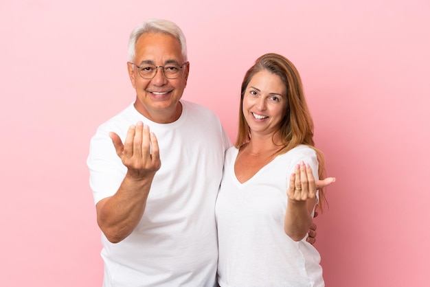 Middle age couple isolated on pink background inviting to come with hand