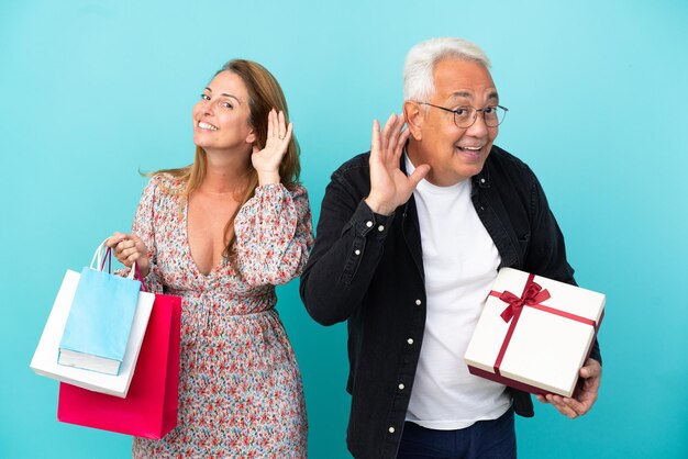 Middle age couple with shopping bag and gift isolated on blue background listening to something by putting hand on the ear