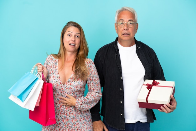 Middle age couple with shopping bag and gift isolated on blue background with surprise and shocked facial expression