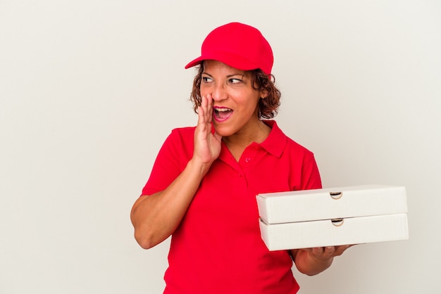 Middle age delivery woman taking pizzas isolated on white background is saying a secret hot braking news and looking aside