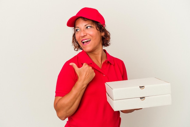 Middle age delivery woman taking pizzas isolated on white background points with thumb finger away, laughing and carefree.