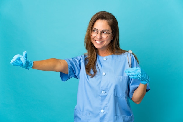 La donna brasiliana del dentista di mezza età che tiene gli strumenti sopra la parete isolata che dà i pollici aumenta il gesto