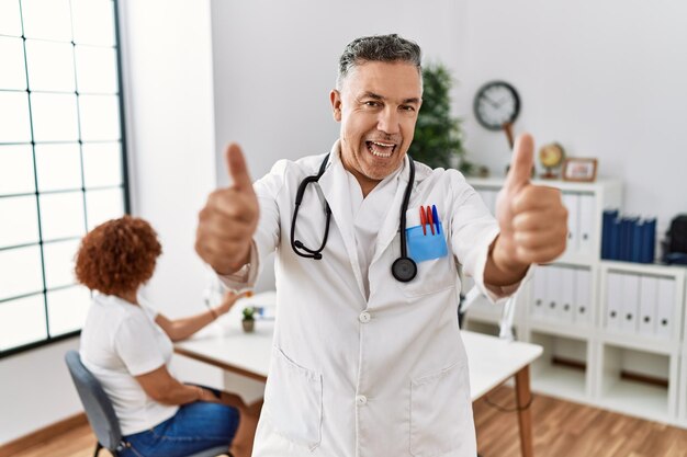 Photo middle age doctor man at the clinic with a patient approving doing positive gesture with hand, thumbs up smiling and happy for success. winner gesture.