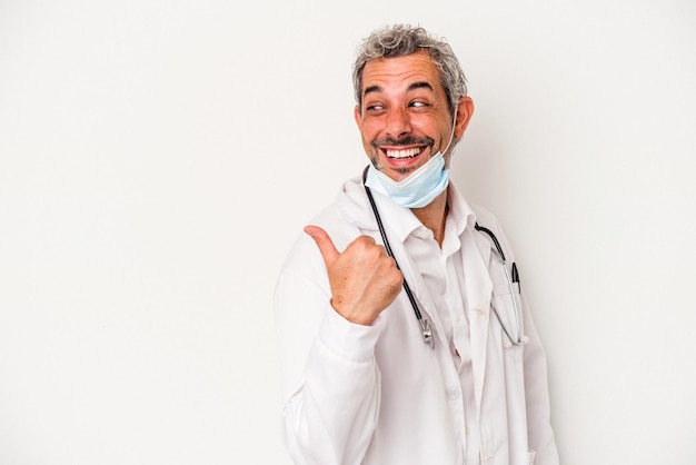 Middle age doctor man wearing a mask for virus isolated on white background  points with thumb finger away, laughing and carefree.