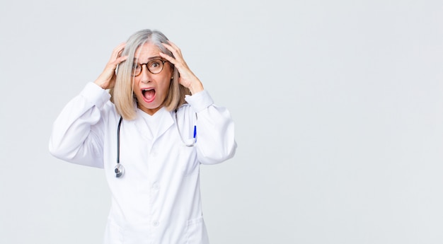 Middle age doctor woman looking excited and surprised, open-mouthed with both hands on head, feeling like a lucky winner