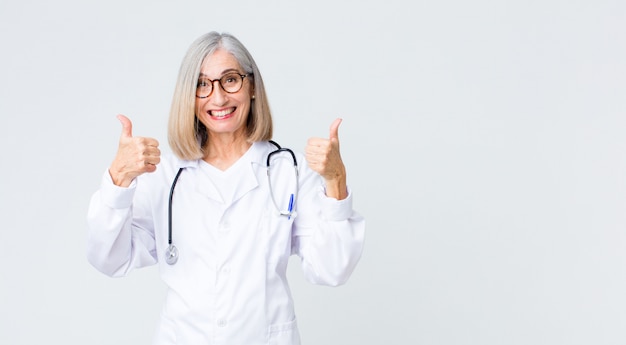 Middle age doctor woman smiling broadly looking happy, positive, confident and successful, with both thumbs up