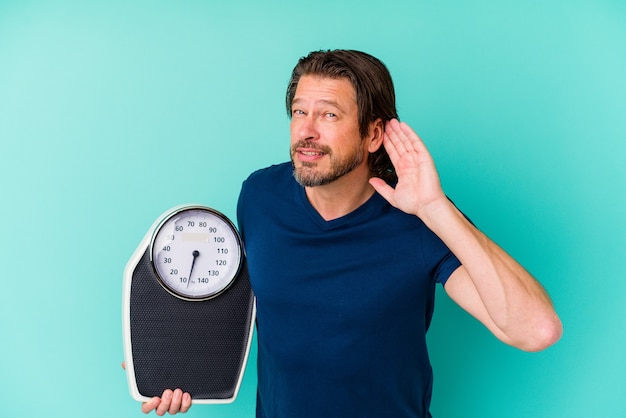 Middle age dutch man holding a scale isolated on blue background trying to listening a gossip.