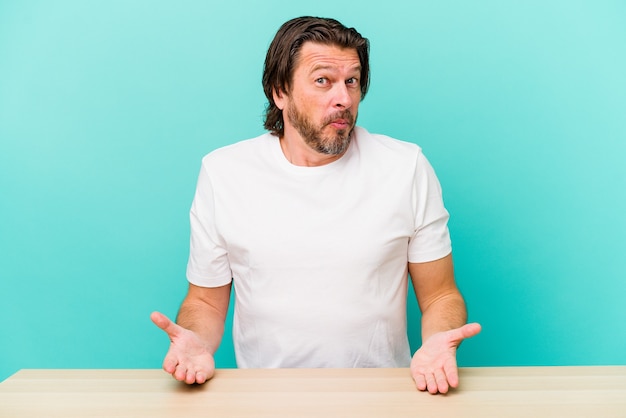 Middle age dutch man sitting isolated on blue background shrugs shoulders and open eyes confused.