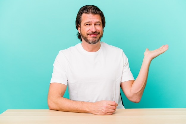 Middle age dutch man sitting isolated on blue wall showing a copy space on a palm and holding another hand on waist.