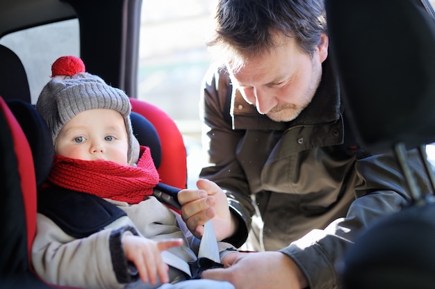 Middle age father helps his toddler son to fasten belt on car seat