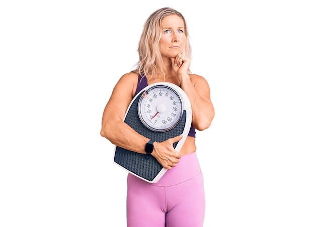 Middle age fit blonde woman wearing sports clothes holding weighing machine serious face thinking about question with hand on chin thoughtful about confusing idea