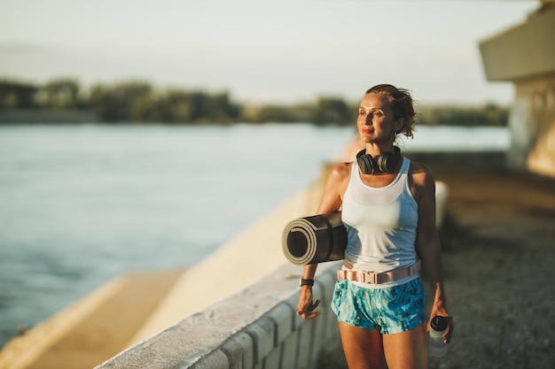 Photo a middle age fit woman holding a yoga mat and water bottle while walking near to river in the city before hard training.