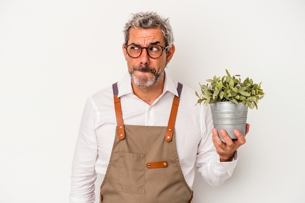 L'uomo caucasico del giardiniere di mezza età che tiene una pianta isolata su fondo bianco confuso, si sente dubbioso e insicuro.