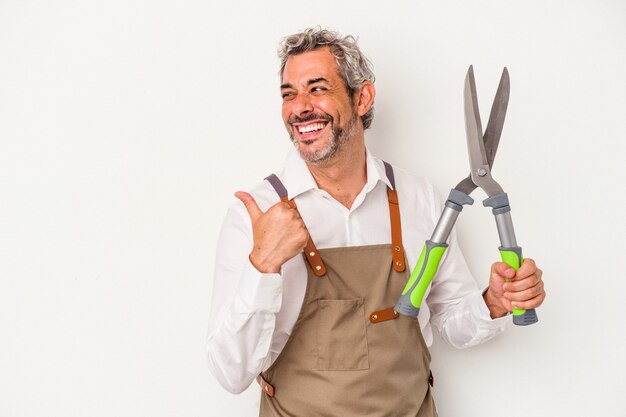 Middle age gardener man holding a scissors isolated on white background  points with thumb finger away, laughing and carefree.