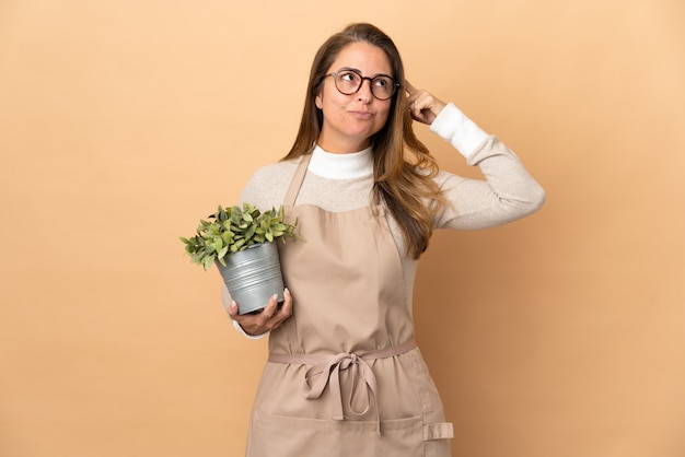 疑問と考えを持っているベージュに分離された植物を保持している中年の庭師の女性