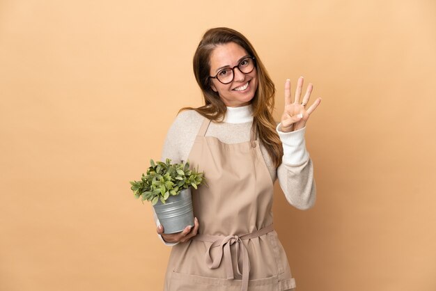 幸せなベージュの壁に隔離された植物を保持し、指で4を数える中年の庭師の女性