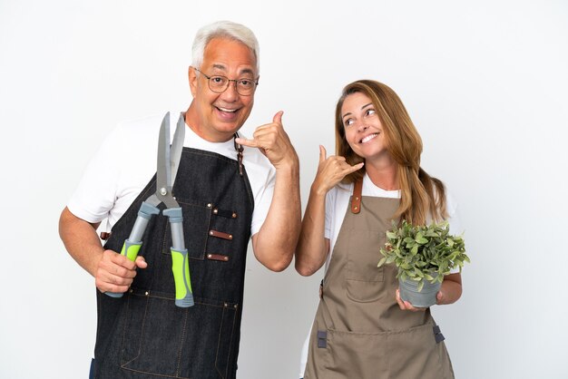 Middle age gardeners holding a plant and scissors isolated on white background making phone gesture. Call me back sign