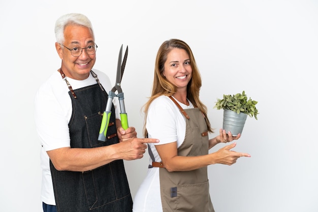 Middle age gardeners holding a plant and scissors isolated on white background pointing finger to the side in lateral position