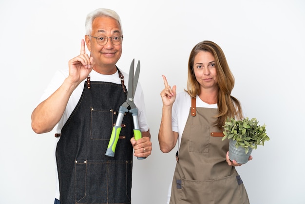 Middle age gardeners holding a plant and scissors isolated on white background showing and lifting a finger in sign of the best