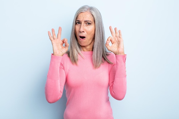 Photo middle age gray hair woman feeling shocked, amazed and surprised, showing approval making okay sign with both hands