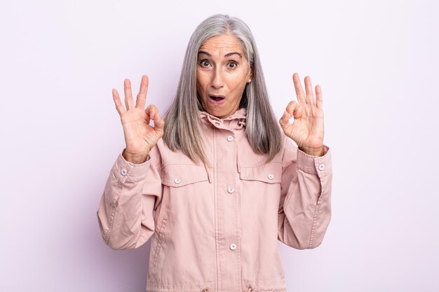 Middle age gray hair woman feeling shocked, amazed and surprised, showing approval making okay sign with both hands