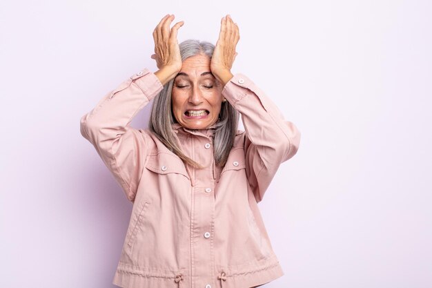 Donna di mezza età con i capelli grigi che si sente stressata e ansiosa, depressa e frustrata per il mal di testa, alzando entrambe le mani alla testa