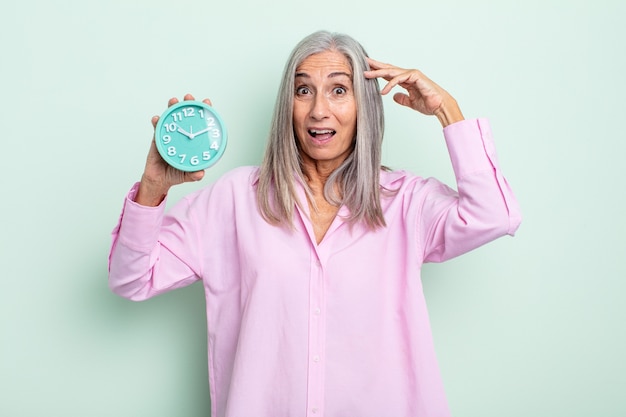 Middle age gray hair woman looking happy, astonished and surprised. alarm clock concept