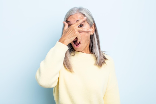 middle age gray hair woman looking shocked, scared or terrified, covering face with hand and peeking between fingers