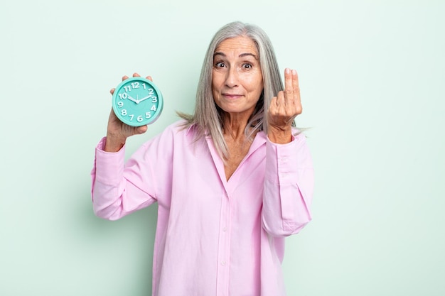 Middle age gray hair woman making capice or money gesture, telling you to pay. alarm clock concept