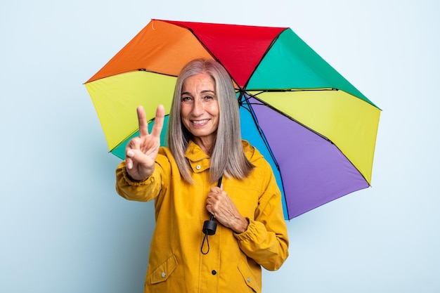 Среднего возраста седые волосы женщина улыбается и выглядит дружелюбно, показывая номер два. зонтик и дождь концепция