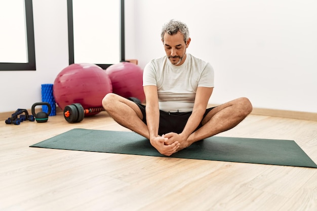 Middle age greyhaired man stretching legs at sport center