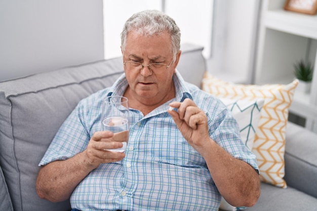 Middle age greyhaired man taking pill drinking glass of water at home