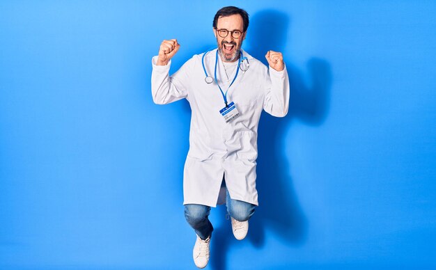 Middle age handsome doctor man wearing stethoscope and coat smiling happy. Jumping with smile on face doing winner gesture with fists up over isolated blue background