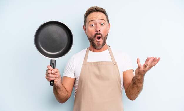 Middle age handsome man amazed, shocked and astonished with an unbelievable surprise. frying pan concept