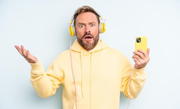 Middle age handsome man feeling extremely shocked and surprised. headphones and smartphone concept